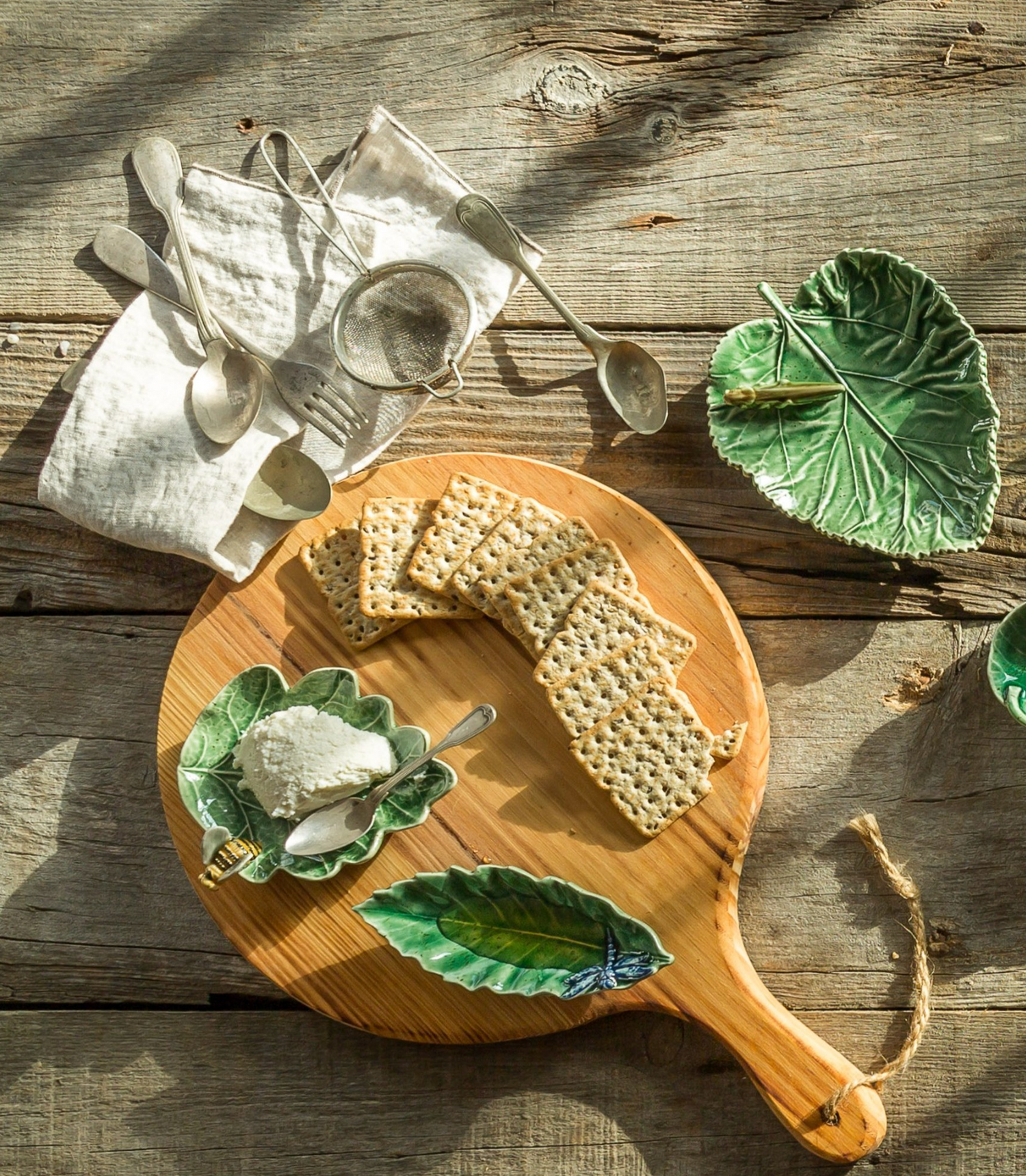 Wooden cutting board with crackers, cheese, and leaf-shaped ceramic plates on a rustic wooden surface.