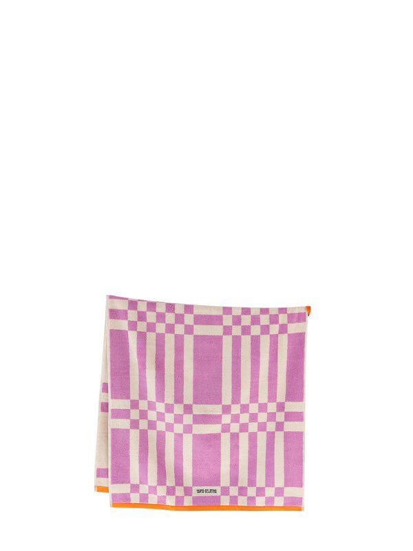 A folded square bath towel featuring a pink and off-white plaid pattern with a thin orange border and a small orange loop at the middle and an off-white Tarta Gelatina brand label at the bottom.