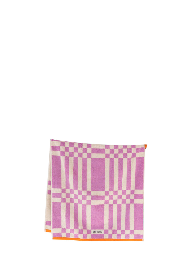 A folded square bath towel featuring a pink and off-white plaid pattern with a thin orange border and a small orange loop at the middle and an off-white Tarta Gelatina brand label at the bottom.