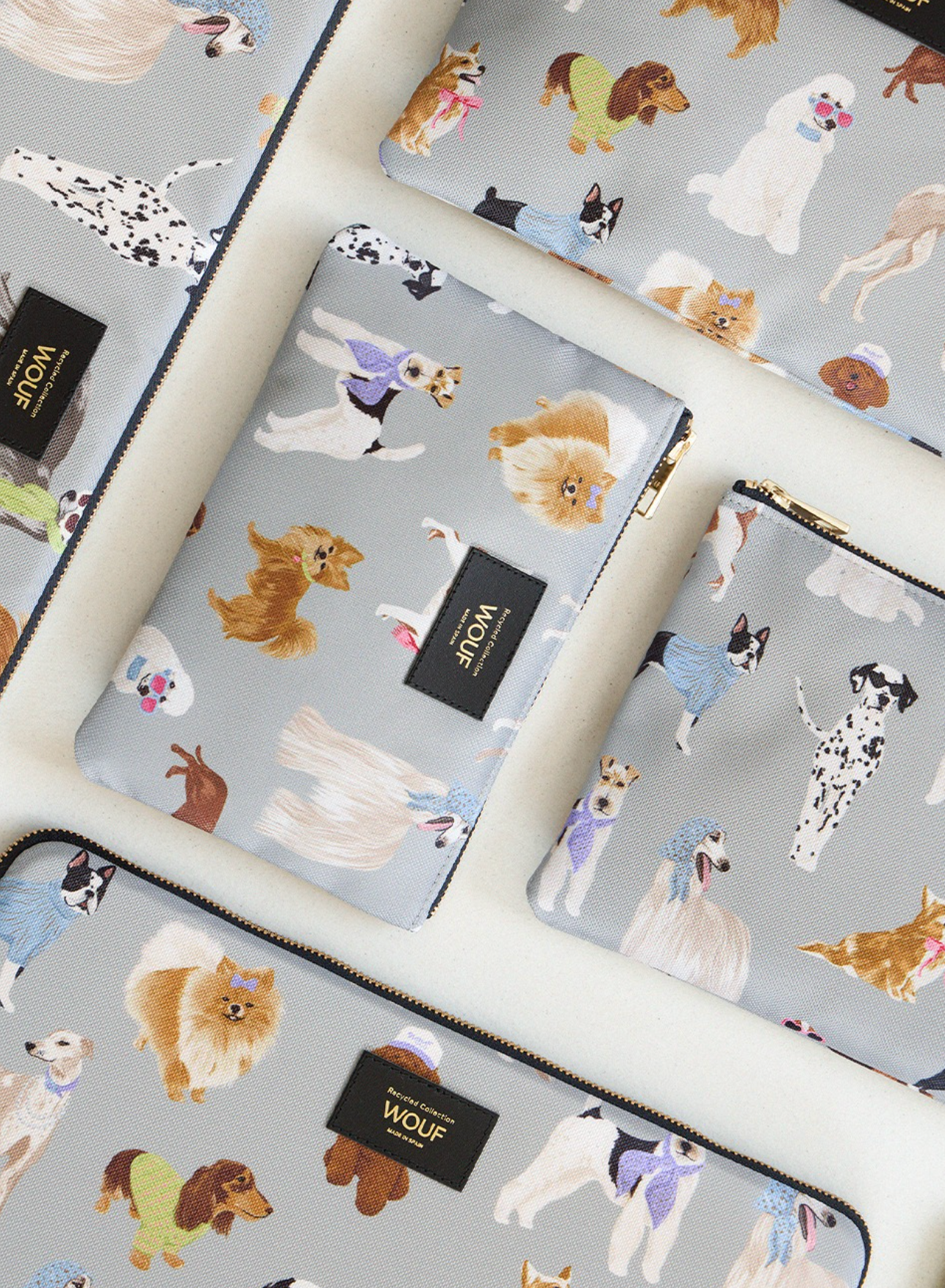 A set of light gray zippered pouches and a matching pouch featuring a colorful cartoon dog print, arranged on a cream-colored surface. Each pouch has a black rectangular brand tab with gold text reading 'WOLF'.