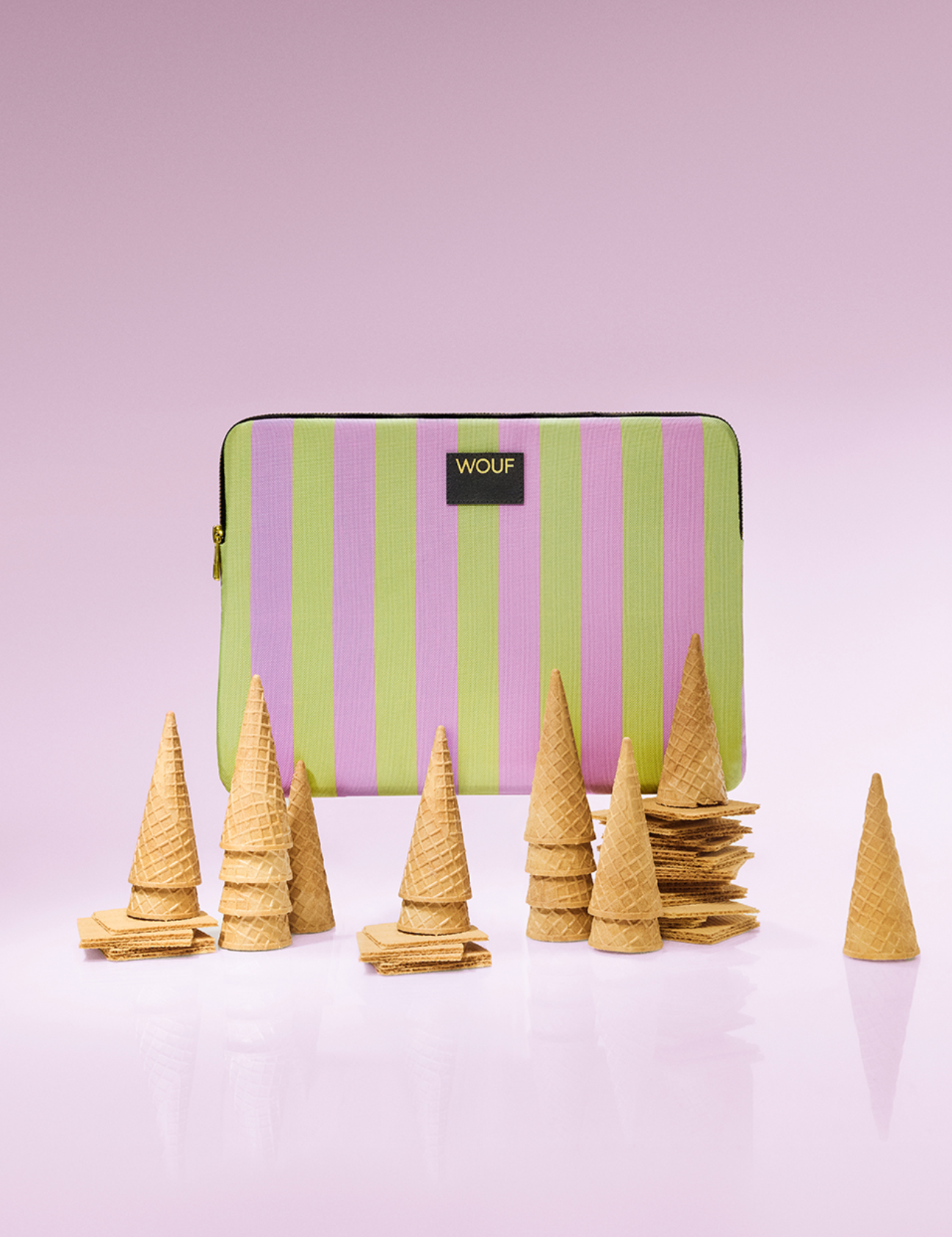 A striped zippered pouch with vertical pink & green stripes, featuring a black label with the brand name 'WOUF'. In front of the pouch are multiple small stacks of waffle cones and waffle pieces arranged on a reflective surface against a solid pink background.