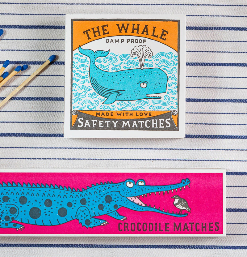 Two matchboxes, one with a whale design and the other with a crocodile design, on a striped background.