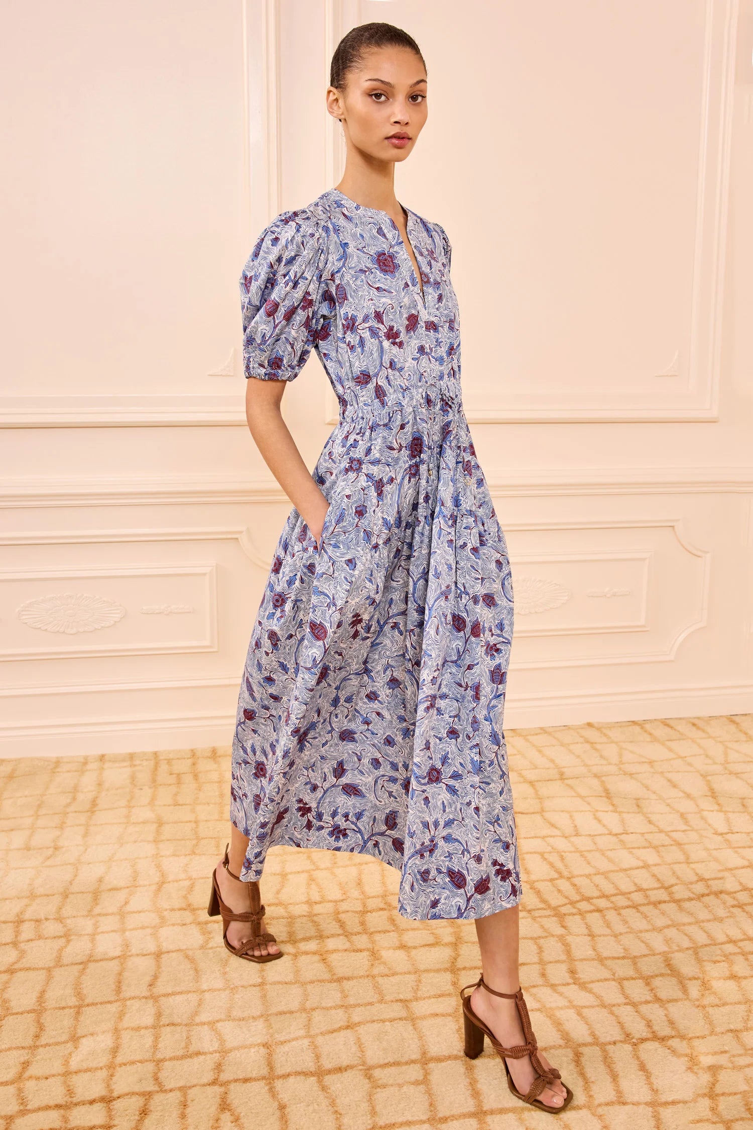 Woman wearing a floral dress standing in a room with a patterned floor and light-colored walls.