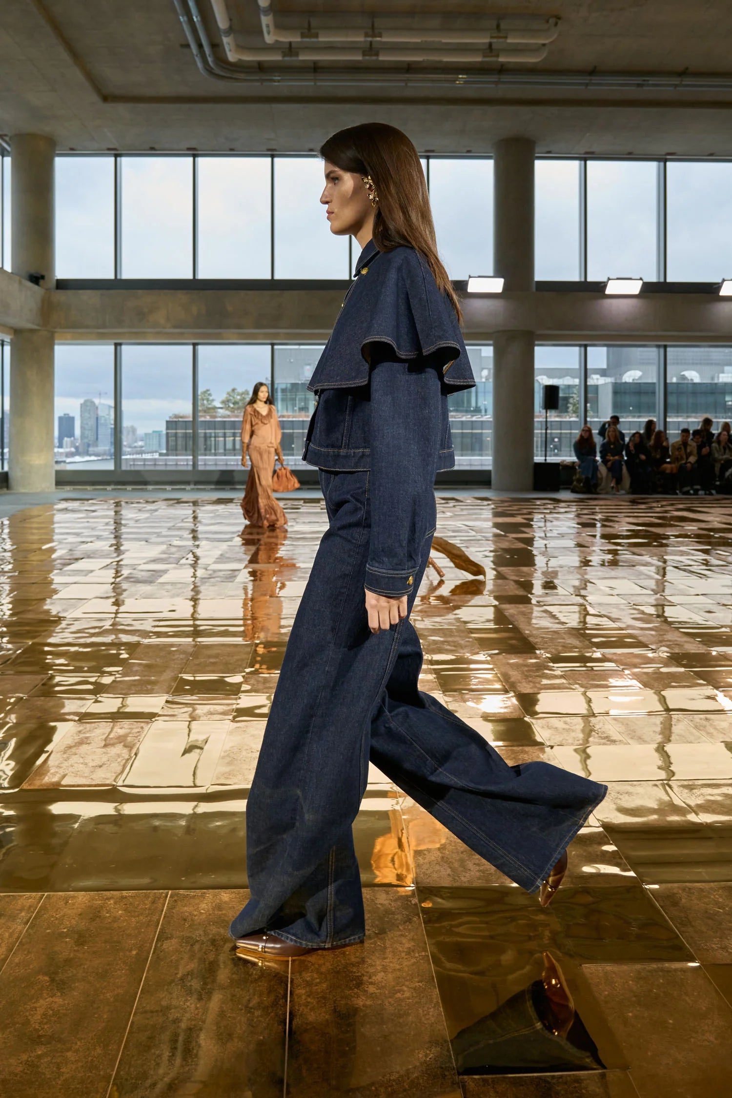 Woman in a blue denim outfit walking in a modern indoor setting with large windows.