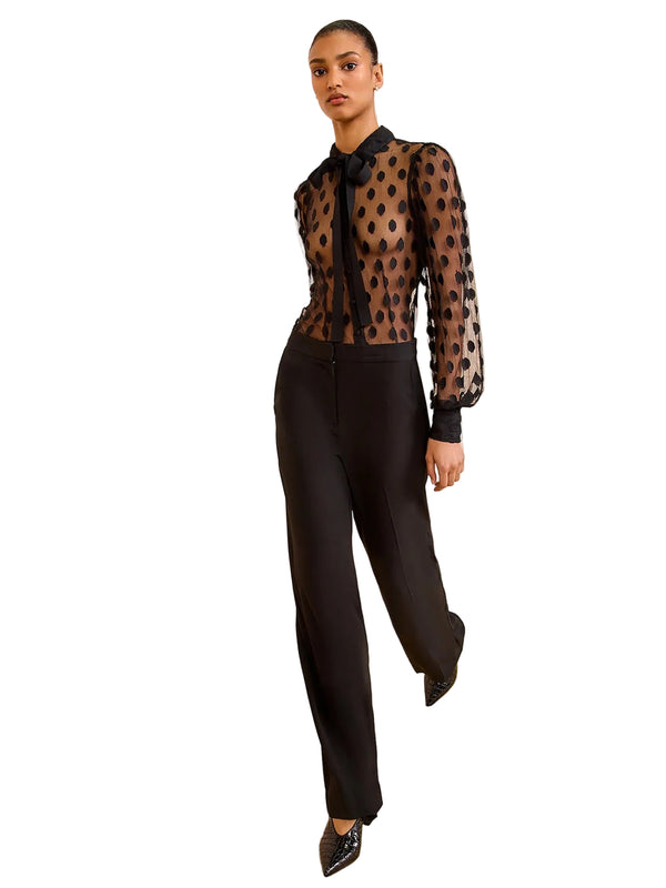 Woman wearing a sheer black polka dot blouse with black pants against a beige wall.