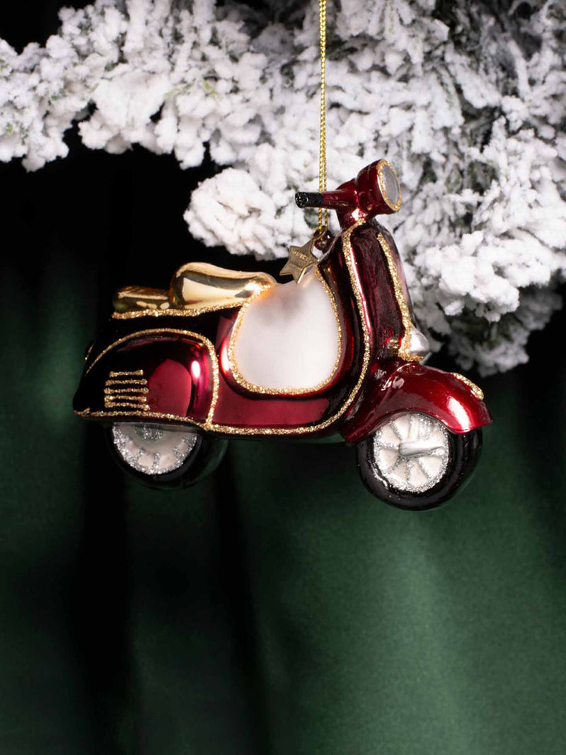 A glossy red and gold scooter-shaped Christmas ornament hanging from a gold string beneath frosted white tree branches against a dark green background.