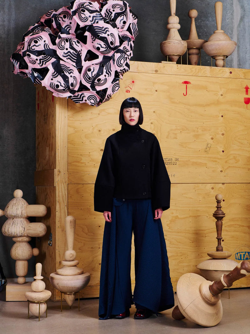 A model stands in a studio setting wearing a black cropped jacket and wide-leg navy trousers, posed in front of a large wooden crate with sculptural wooden objects around them and a pink patterned umbrella overhead.