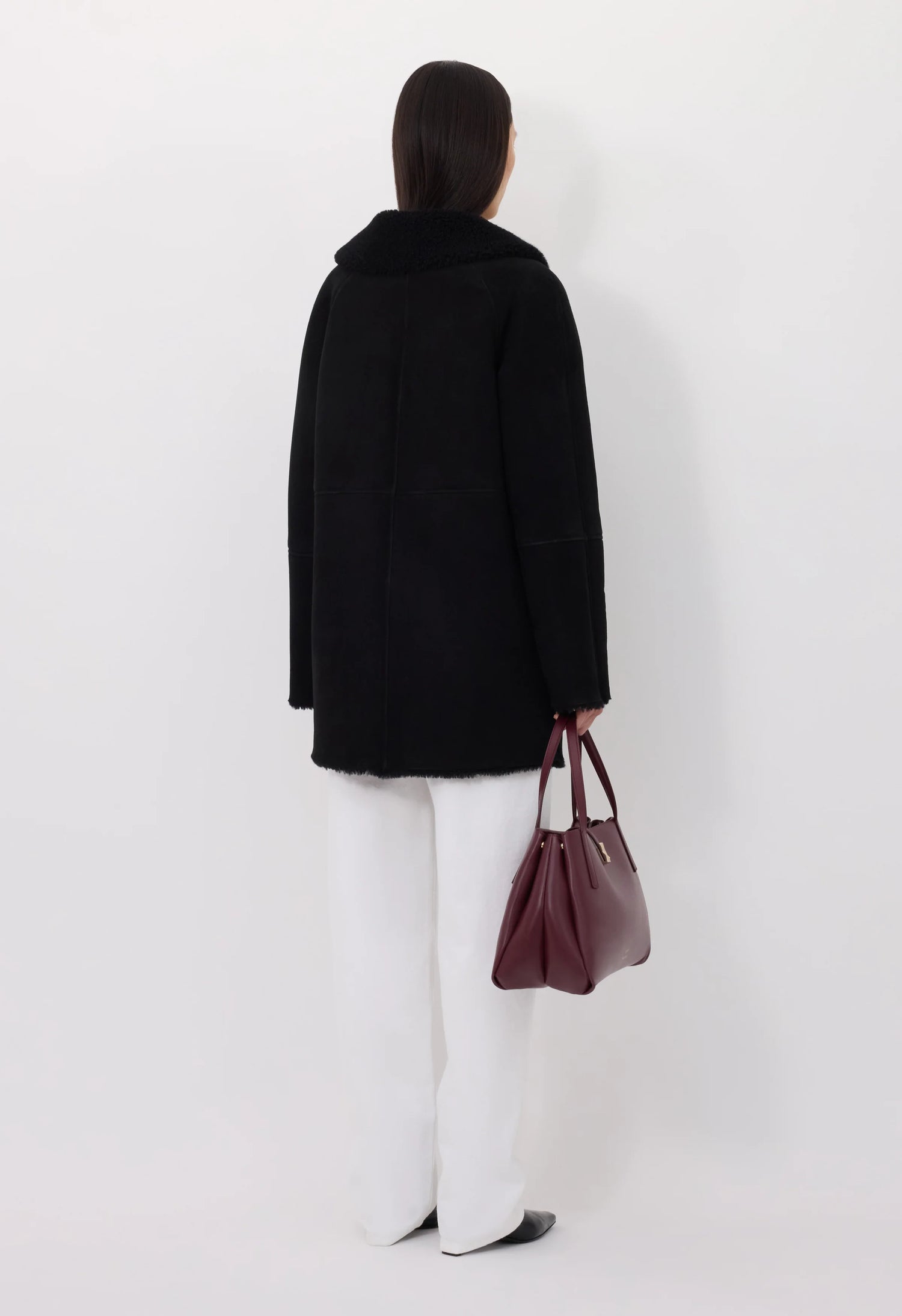 Person wearing a black shearling coat and white pants holding a brown bag against a white background