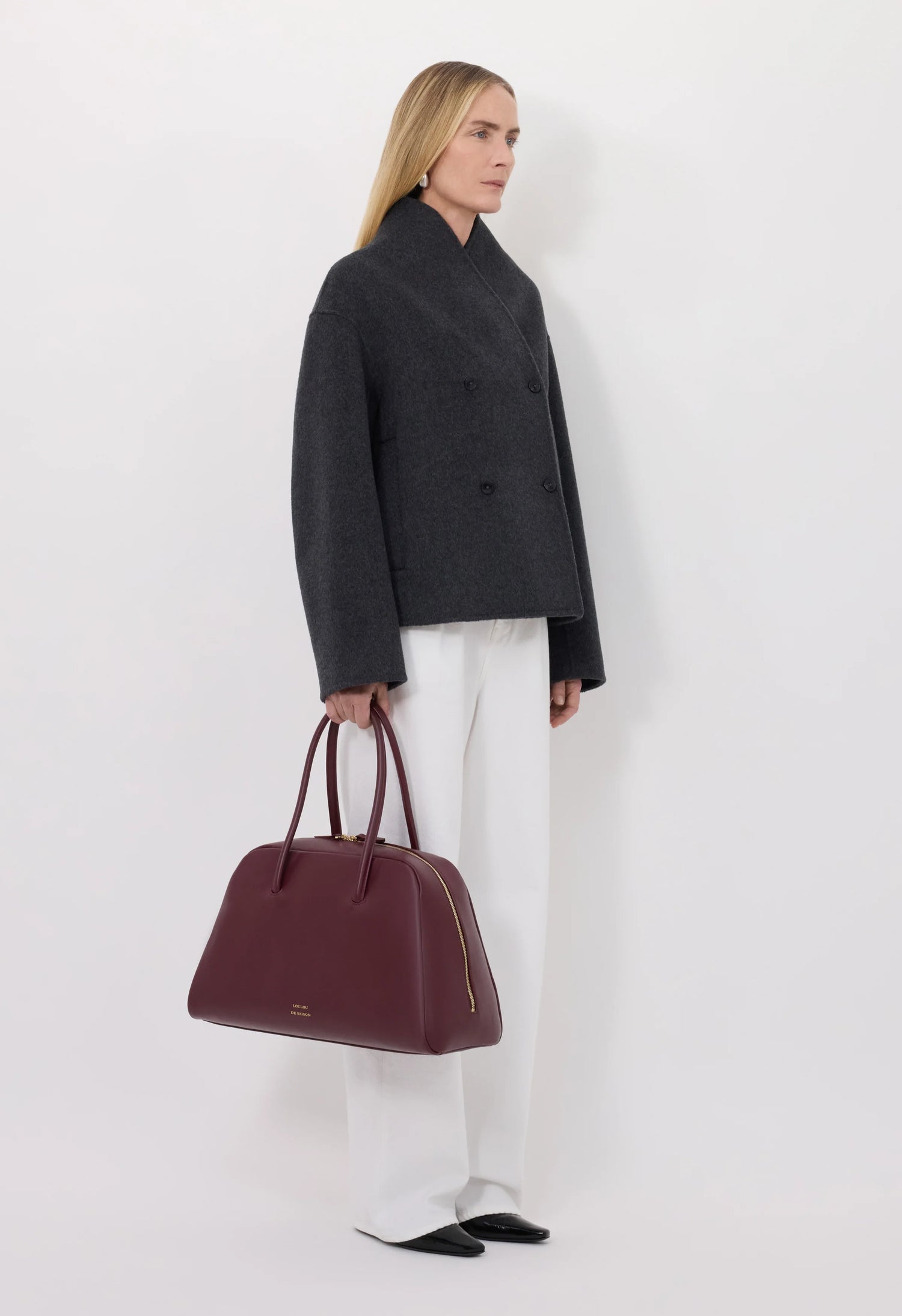Person wearing a dark gray coat and white pants, holding a burgundy handbag against a white background