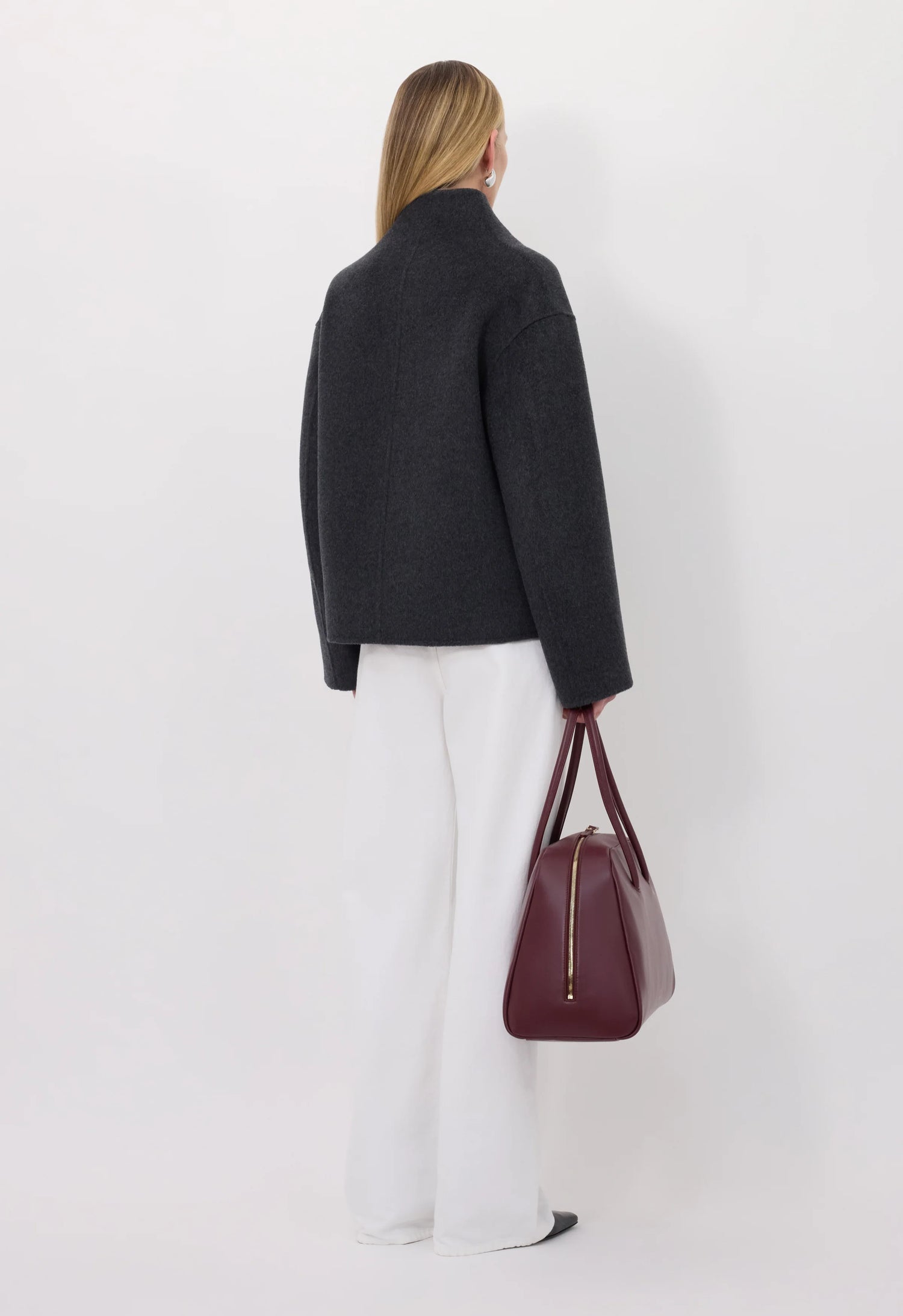 Person wearing a grey coat and white pants holding a maroon handbag on a white background