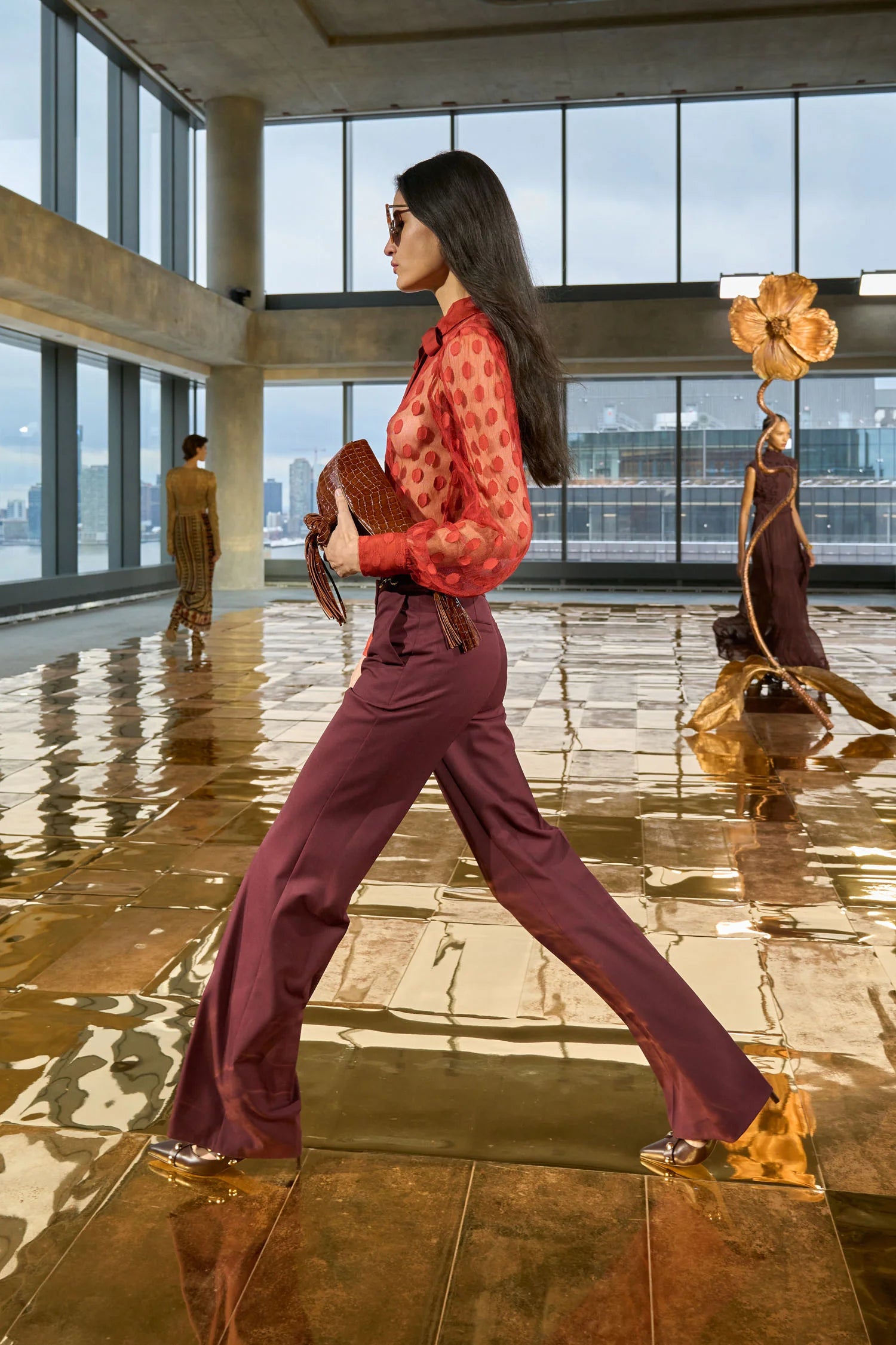Woman in a red blouse and purple pants walking in a modern building with large windows.