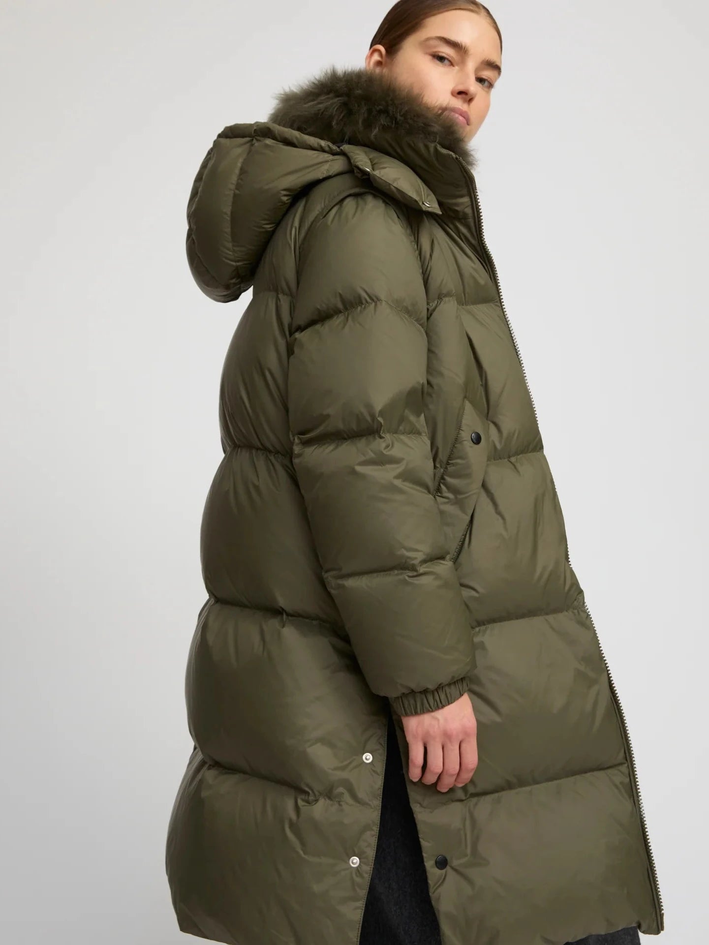Person wearing a long green puffer coat with a fur-lined hood on a light gray background