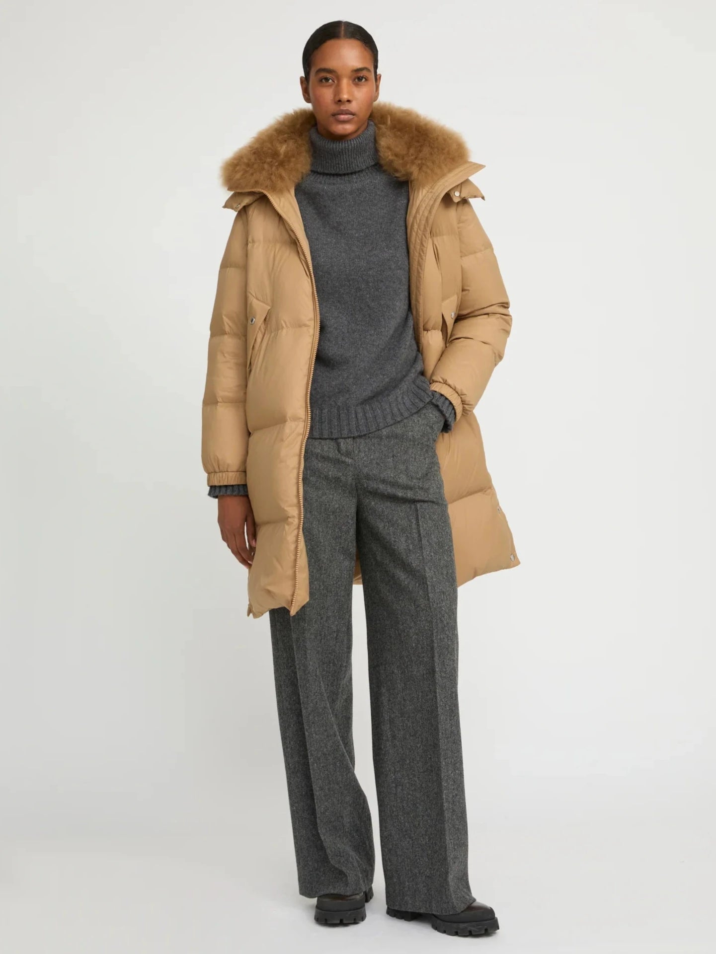 Person wearing a beige puffer coat with a fur-lined hood and gray sweater and pants on a white background
