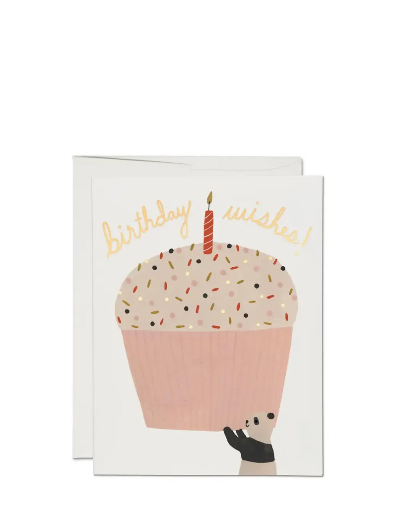 A birthday card with an illustration of a cupcake with sprinkles and a candle, with the text 'birthday wishes!' on top. The card is pink and there is a white envelope included.
